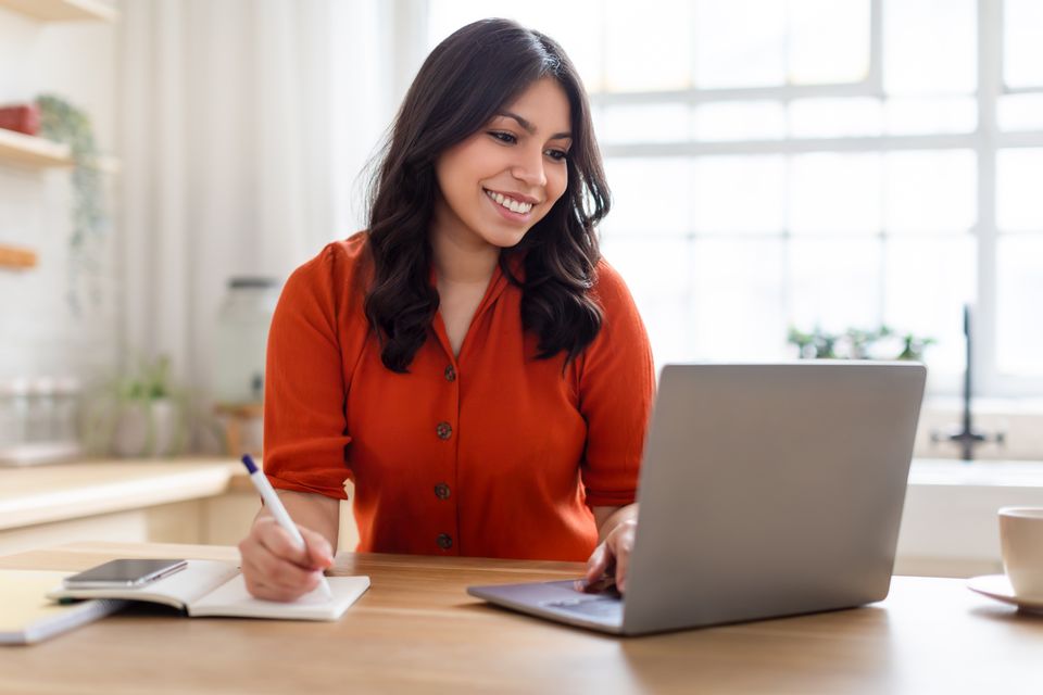 Mujer sonriente toma notas mientras crea su sitio web en una laptop en la cocina de su casa